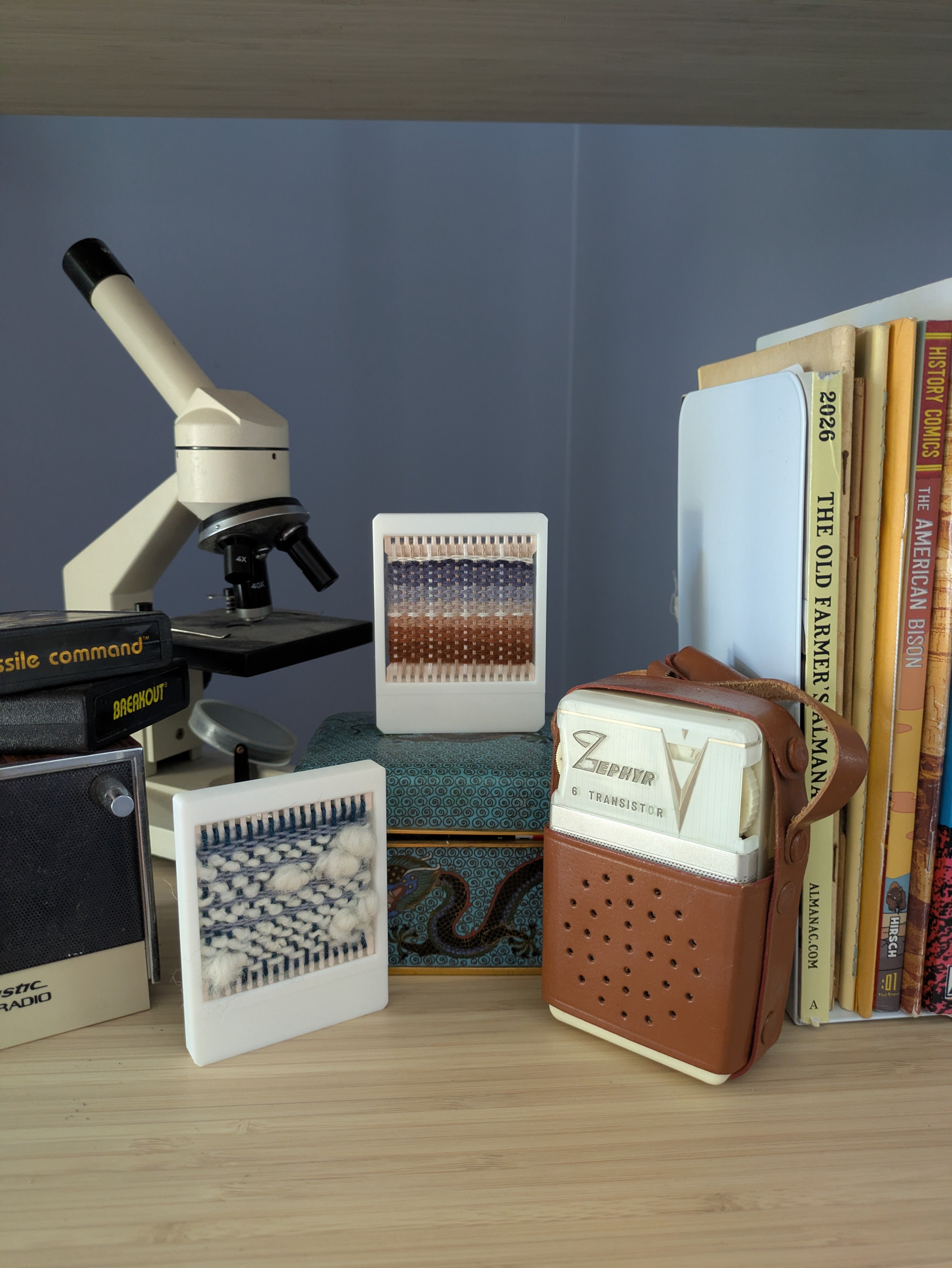 Two swatch loom retro rames with a vintage-style brown leather and cream plastic speaker-shaped bookend with a brown handle, displayed on a light wood shelf next to books, a microscope, and patterned tiles against a blue-gray wall.